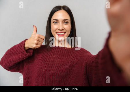 Photo de jolie jeune femme vêtue de chandail bourgogne prendre en selfies isolés de l'appareil photo sur fond de mur gris. Banque D'Images