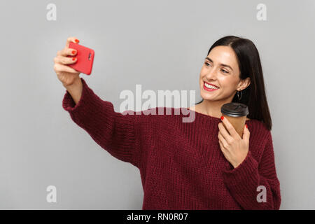 Photo de jolie jeune femme vêtue de chandail bourgogne selfies prendre par téléphone mobile sur fond de mur gris isolé de boire du café. Banque D'Images