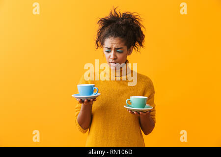 Confus african woman wearing sweater holding tasses soucoupes plus isolé sur fond jaune Banque D'Images