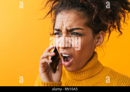 Close up of a choqué confus african woman wearing sweater isolés sur fond jaune, talking on mobile phone Banque D'Images