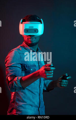 Jeune homme africain méconnaissable playing video game portant la réalité virtuelle contre l'appareil à deux couleurs rouge et bleu sur fond noir. Des jeux de l'eq Banque D'Images