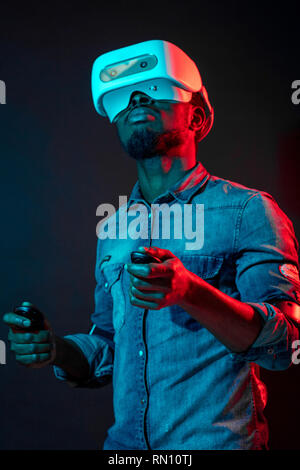 Jeune homme africain méconnaissable playing video game portant la réalité virtuelle contre l'appareil à deux couleurs rouge et bleu sur fond noir. Des jeux de l'eq Banque D'Images