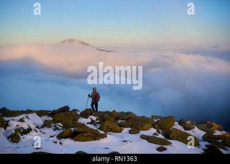 Vue arrière de l'randonneur avec sac à dos et bâtons randonnée sur rocky hill sur fond de vallée brumeuse magnifique rempli de puffy blanc nuages, montagne enneigée haut et ciel bleu le matin. Banque D'Images