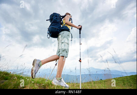 Low angle view of attractive woman randonneur randonnée en sentier de montagne des Carpates, balade sur la colline herbeuse, le port de sac à dos, à l'aide de bâtons de trekking, profitant de journée d'été. L'activité de plein air, style de concept Banque D'Images