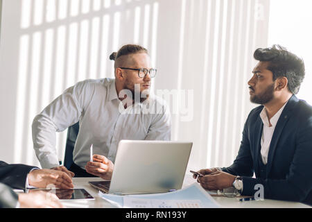 Homme d'optimisme confiant en chemise blanche parler avec son subalterne indien ou assistant en étant assis dans une réunion informelle dans office Banque D'Images