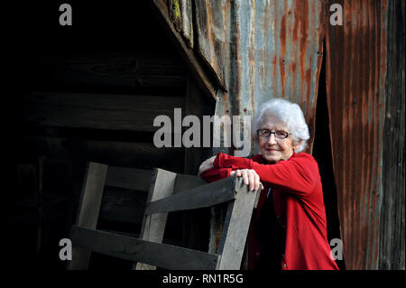 Femme âgée s'appuie contre une partie de l'ancienne grange familiale en Arkansas. Elle a les cheveux blancs et porte des lunettes. Elle porte une veste rouge. Banque D'Images