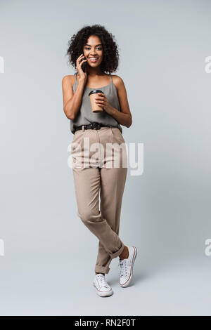 Toute la longueur d'une jeune femme africaine de désinvolture habillé isolés sur fond gris,talking on mobile phone, holding cup à emporter de co Banque D'Images