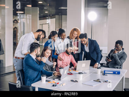 L'interracial deux gars dans le travail de bureau usure formelle jouer jeu en ligne sur les ordinateurs portables pendant que leurs collègues joyeux excité réunir autour de la table supp Banque D'Images