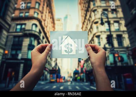 La pauvreté et l'itinérance enjeu global concept comme femme mains tenant une feuille de papier avec la maison et l'intérieur de l'icône du dollar au milieu de la rue d'un Banque D'Images