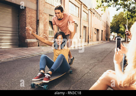 Couple sur planche avec les amis de prendre leurs photos. Man pushing woman on skateboard avec leurs amis assis par la route prendre thei Banque D'Images