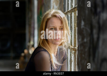 Belle fille blonde avec des yeux verts et des cheveux ébouriffés longue fixant songeur à l'appareil photo avec parted lèvres aux côtés d'une vieille fenêtre à l'intérieur rustique Banque D'Images