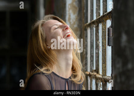 Heureux amusé jeune femme bénéficiant d'un bon rire avec sa tête rejetée en arrière et fermé les yeux devant une grande fenêtre à l'intérieur rustique Banque D'Images