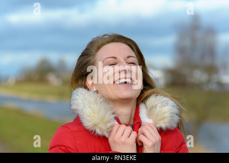 Jeune femme joyeuse en fourrure manteau d'hiver parés à l'extérieur permanent dans la campagne de rire avec sa tête rejetée en arrière et les yeux fermés Banque D'Images