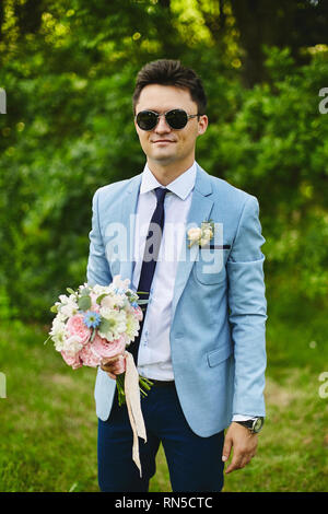 Beau jeune groom dans le quartier à la veste bleue avec une cravate et des lunettes de soleil élégantes avec le bouquet de fleurs dans ses mains Banque D'Images