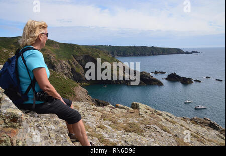 Femme seule randonneur assis sur les falaises à la recherche sur les bateaux du port de Le Havre Gosselin depuis le sentier du Littoral sur l'île de Sark, Channel Islands, Royaume-Uni. Banque D'Images