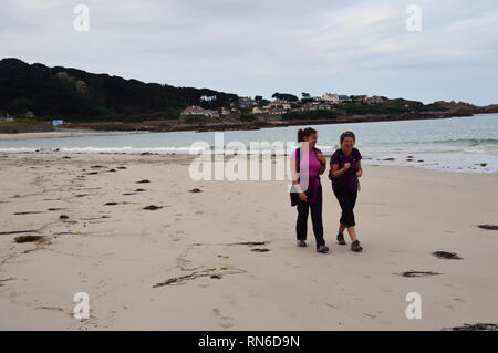 Deux femmes randonneurs marcher, parler et rire sur Cobo Bay Beach Guernsey, Channel Islands. UK. Banque D'Images