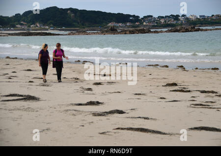 Deux femmes randonneurs marcher, parler et rire sur Cobo Bay Beach Guernsey, Channel Islands. UK. Banque D'Images