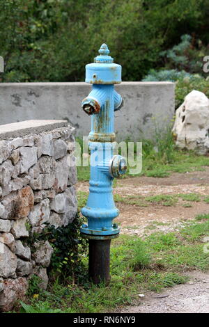 Vintage ancien rouillé métal bleu monté sur poteau incendie tuyau métallique solide à côté de mur de pierre traditionnel entouré d'arbres et l'herbe non coupée Banque D'Images