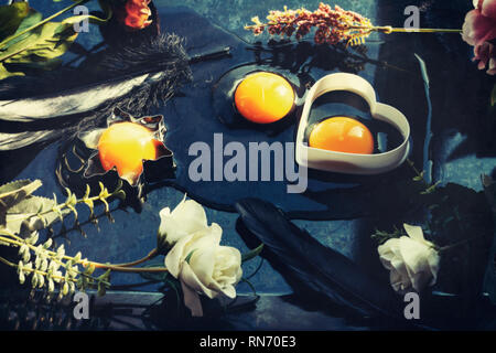 Trois oeufs crus blancs et jaunes, deux dans des emporte-pièce, l'un renversé sur une table, décoré de fleurs de printemps et plumes noires pour les vacances de Pâques Banque D'Images