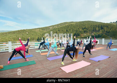 Pose d'tournait triangle. Les gens de la pratique de groupe yoga asana, jusqu'à maintenant, jusqu'à la main. Les femmes sportives dans les vêtements de sport, de détente en plein air. Belle vue sur montagnes et ciel bleu. Banque D'Images