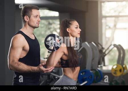 Personnel professionnel trainer helping woman in gym avec des exercices. La belle brunette barbell, entraîneur en charge. Charmante femme aux cheveux longs portant en noir haut sportive. Banque D'Images