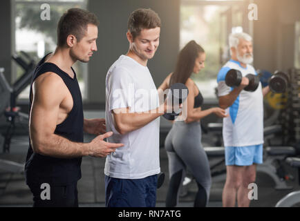 Sportifs l'entraînement en salle de sport avec des entraîneurs personnels. Coach musculaire en noir sleeveless sports shirt homme aidant à faire les exercices. Entraîneur féminin debout près de vieil homme. Banque D'Images