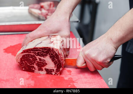 Libre de main du cuisinier coupe Ribeye Steak de boeuf persillé avec un couteau sur une planche à découper en plastique rouge sur métal table dans une cuisine de restaurant. Concept steakho Banque D'Images