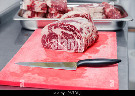 Pièces macro closeup Ribeye Steak de boeuf marbré au couteau sur une planche à découper en plastique rouge sur métal table dans une cuisine de restaurant. Concept emballage semi-fini Banque D'Images