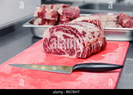 Pièces macro closeup Ribeye Steak de boeuf marbré au couteau sur une planche à découper en plastique rouge sur métal table dans une cuisine de restaurant. Concept emballage semi-fini Banque D'Images
