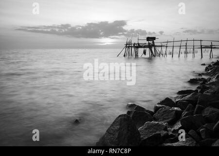 Jetée en bois avec le bord de mer rocheux pendant le coucher du soleil. Une longue exposition noir et blanc Banque D'Images