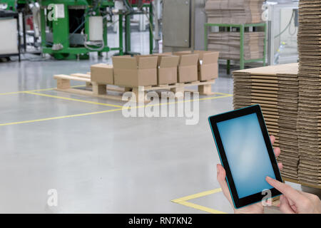 Hall de production vide avec des boîtes en papier plié sur des palettes en bois et les mains tenant tablette avec écran blanc prêt pour votre texte. Banque D'Images