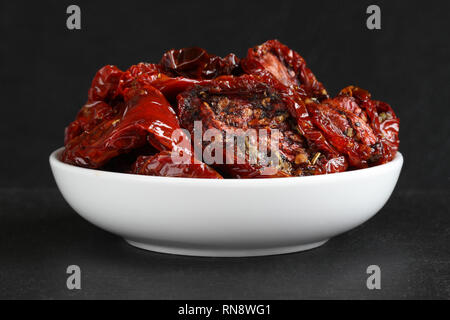 Tomates séchées dans l'huile de tournesol avec des épices (romarin, ail, basilic, poivre noir) dans un bol en céramique blanc, debout sur une pierre noire aga de surface Banque D'Images