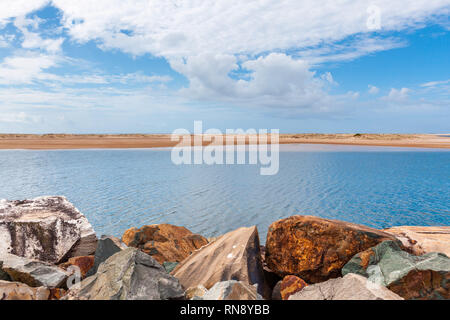 Pierres de brise-lames sur eaux calmes et étendue de sable sous un ciel bleu - paysage minimaliste Banque D'Images