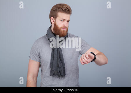 Beau jeune homme barbu wearing scarf debout près du mur gris regardant sa montre le contrôle de temps. Banque D'Images