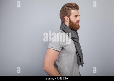 Beau jeune homme barbu debout près du mur gris profil tourné dans le port de foulard et un t-shirt gris. Banque D'Images