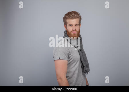 Beau jeune homme barbu debout près du mur gris wearing scarf et t-shirt gris tourné de profil avec la tête tournée vers l'appareil photo. Banque D'Images