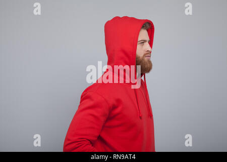 Beau jeune homme barbu tourné de profil portant des sweat rouge debout à l'arrière-plan gris regardant droit devant lui. Banque D'Images