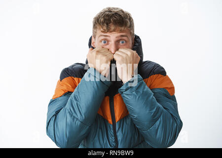 Prêt pour l'hiver. Peur bon-à'homme blond aux yeux bleus se tirant chaud col de puffer jacket sur le visage sur l'expiration de l'air, Comité permanent Banque D'Images