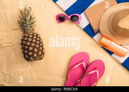 Vue de dessus accessoires d'été, écran solaire sur une serviette à rayures et d'ananas sur le sable Banque D'Images