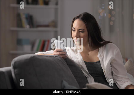 Femme sérieuse contrôle contenu smartphone assis sur un canapé dans la nuit à la maison Banque D'Images