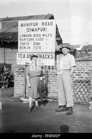 Un moyen âge européen blanc homme et femme posant devant un panneau indiquant le Manipur Road à Jodhpur, Inde. Manipur Road à Jodhpur (État d'Nāgāland) est une ville de l'Inde sur 1 033 km (1 663 km) ou l'Est de New Delhi, la capitale du pays, ville. Vintage milieu xxe siècle photographie noir et blanc. Distances donne signe de diverses villes à travers le monde. Banque D'Images