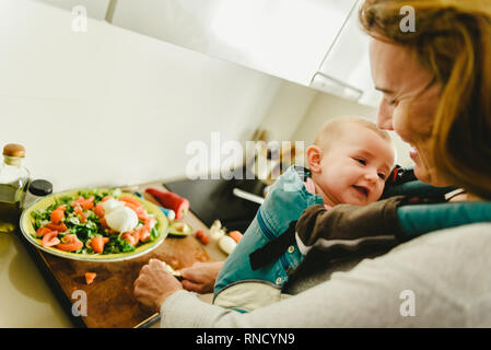 Smiling baby porté dans un porte-bébé sac à dos à la recherche de sa mère alors qu'elle cuisine, concept de conciliation familiale Banque D'Images