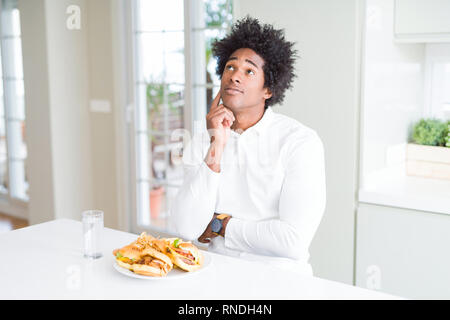 African American hungry man eating hamburger pour le déjeuner avec la main sur le menton de penser à la question, l'expression pensive. Smiling avec visage. N Banque D'Images