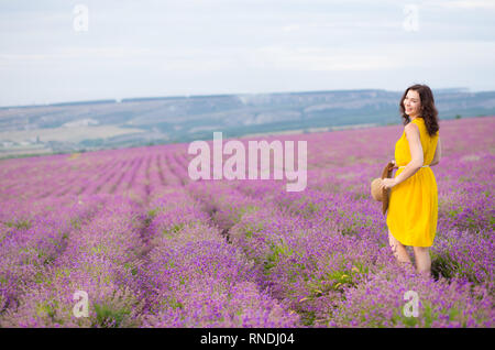 Belle jeune femme en robe jaune avec un chapeau dans les mains se détendre et s'amuser sur champ de lavande fleurs de mauve. Banque D'Images