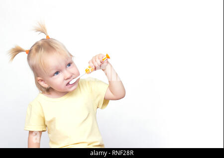 La petite fille la blonde avec des nattes sur la tête de l'apparence caucasienne brosse les dents et regarde ailleurs, fond blanc, copy space, happi Banque D'Images