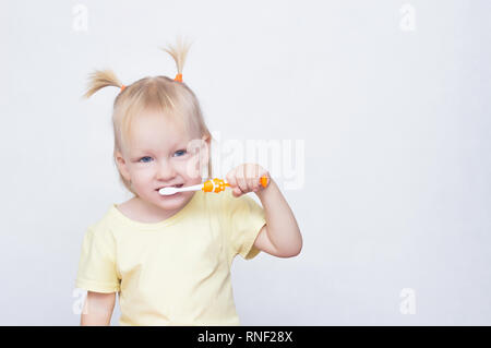 Little blue-eyed girl blonde avec des tresses sur sa tête se brosser les dents avec une brosse à dents et regardant la caméra, de jeunes, de race blanche, l'espace de copie Banque D'Images