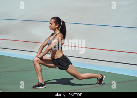 Jeune femme portant des vêtements noirs qui s'étend sur racetrack au cours de session de formation. Coureuse pratiquer l'athlétisme sur piste de course. Elle est concentrée et motivée. Objectifs du corps. Vue de côté Banque D'Images