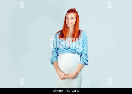 Jolie femme adorable avec redhead hairstyle embracing pregnant belly en se tenant sur le fond bleu clair et donner sourire à la caméra. Banque D'Images