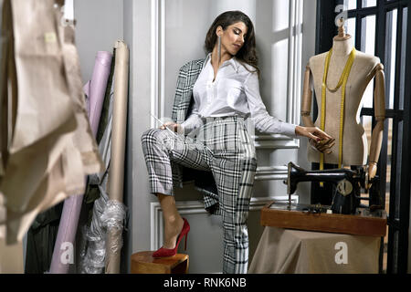 Créateur de mode attrayant est titulaire d'un crayon et d'une main d'un mannequin dans l'atelier avec machine à coudre et des rouleaux de tissu. Elle porte un damier gris s Banque D'Images