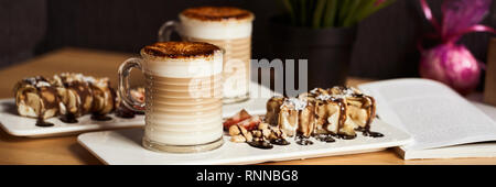 Taille de la bannière image de deux tasses de café au lait chaud avec croûte de caramel cuit et sucré avec des rouleaux de banane et de fraise sur la table en bois dans la région de coffe-shop. Coffe Banque D'Images